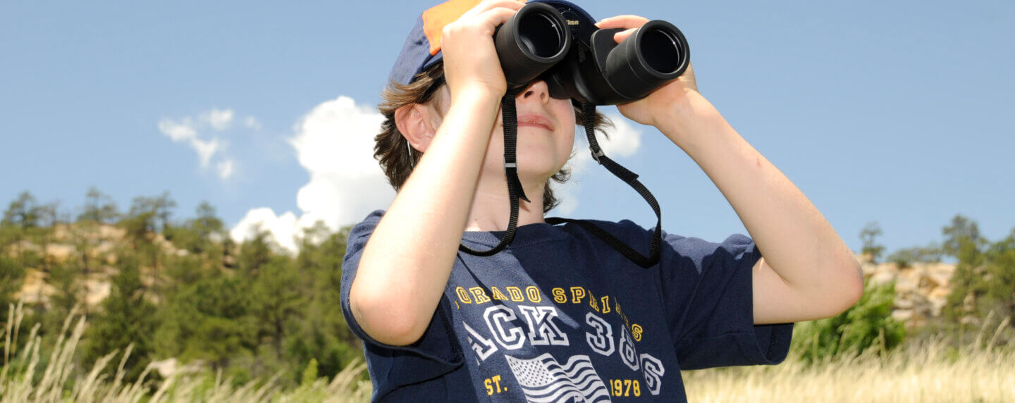 Cub scout standing in a field looking through binoculars